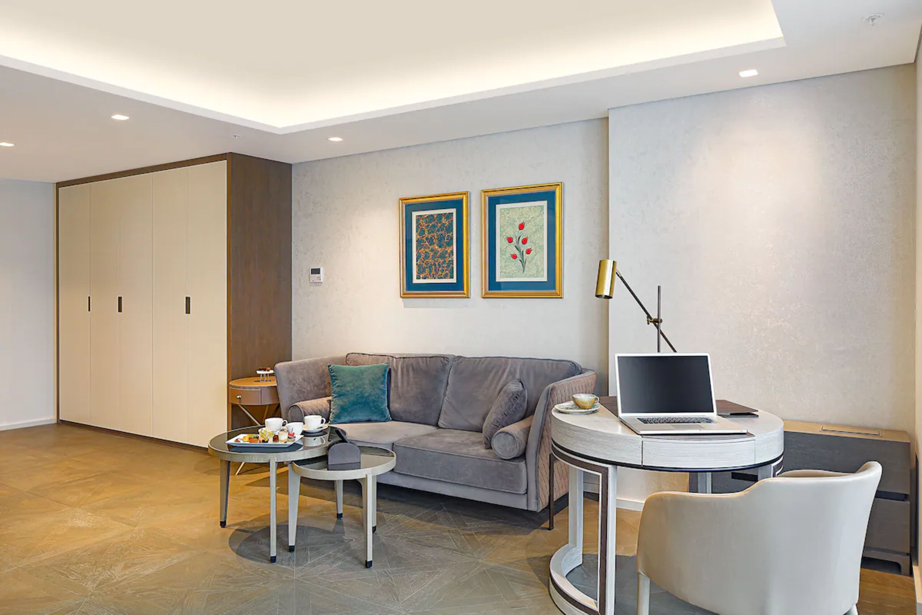 Een moderne woonkamer met een grijze bank, een ronde salontafel, een laptop op een bijzettafeltje en twee ingelijste kunstwerken aan de muur.