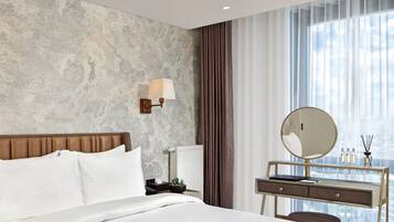 A hotel room with a bed, a desk with a mirror, a lamp, and a view of the sky through sheer curtains.