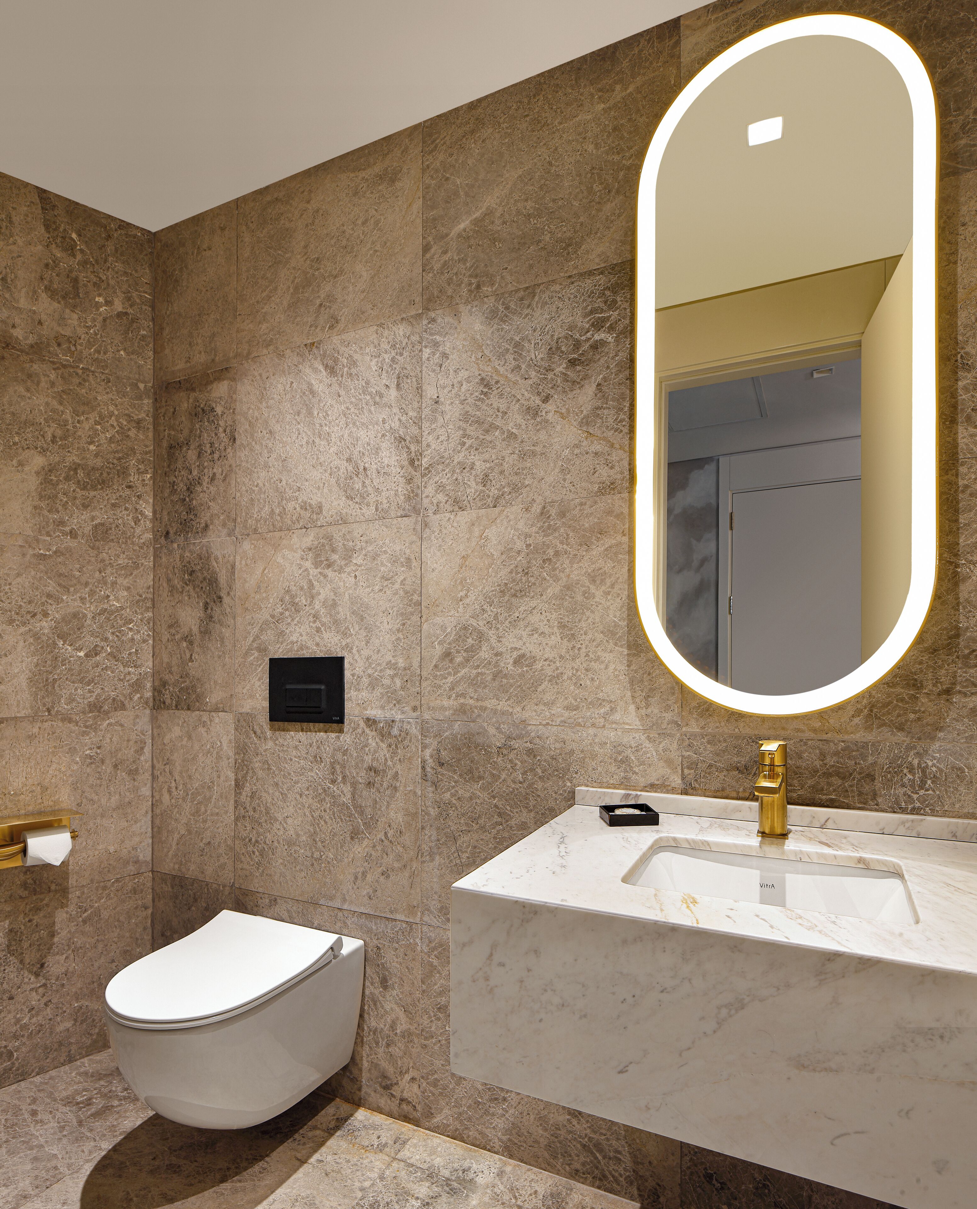 A modern bathroom with a marble wall, a toilet, a sink with a gold faucet, and a mirror with a lighted frame.