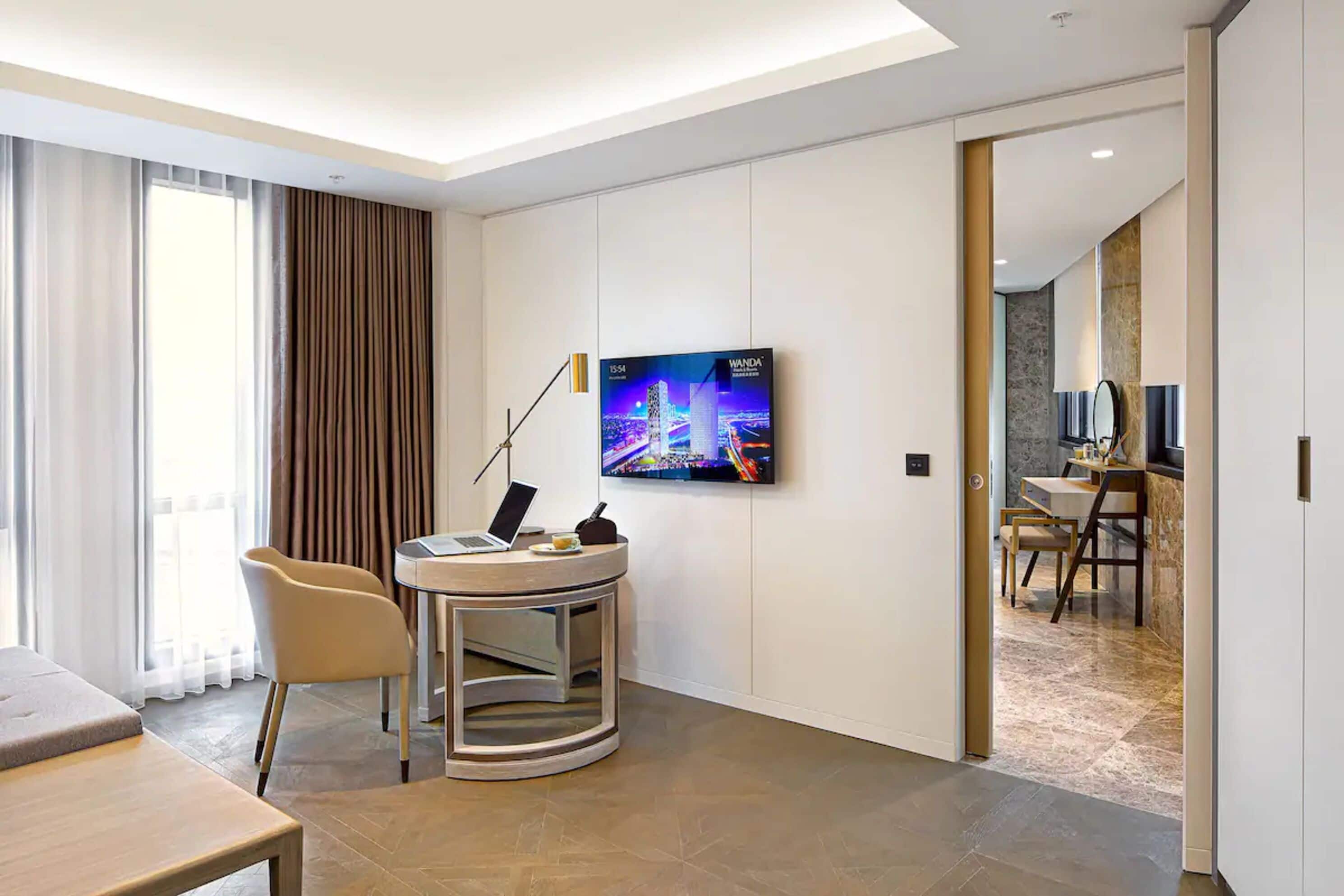 A modern hotel room with a round table, a chair, a TV mounted on the wall, and a dining area visible through an open door.
