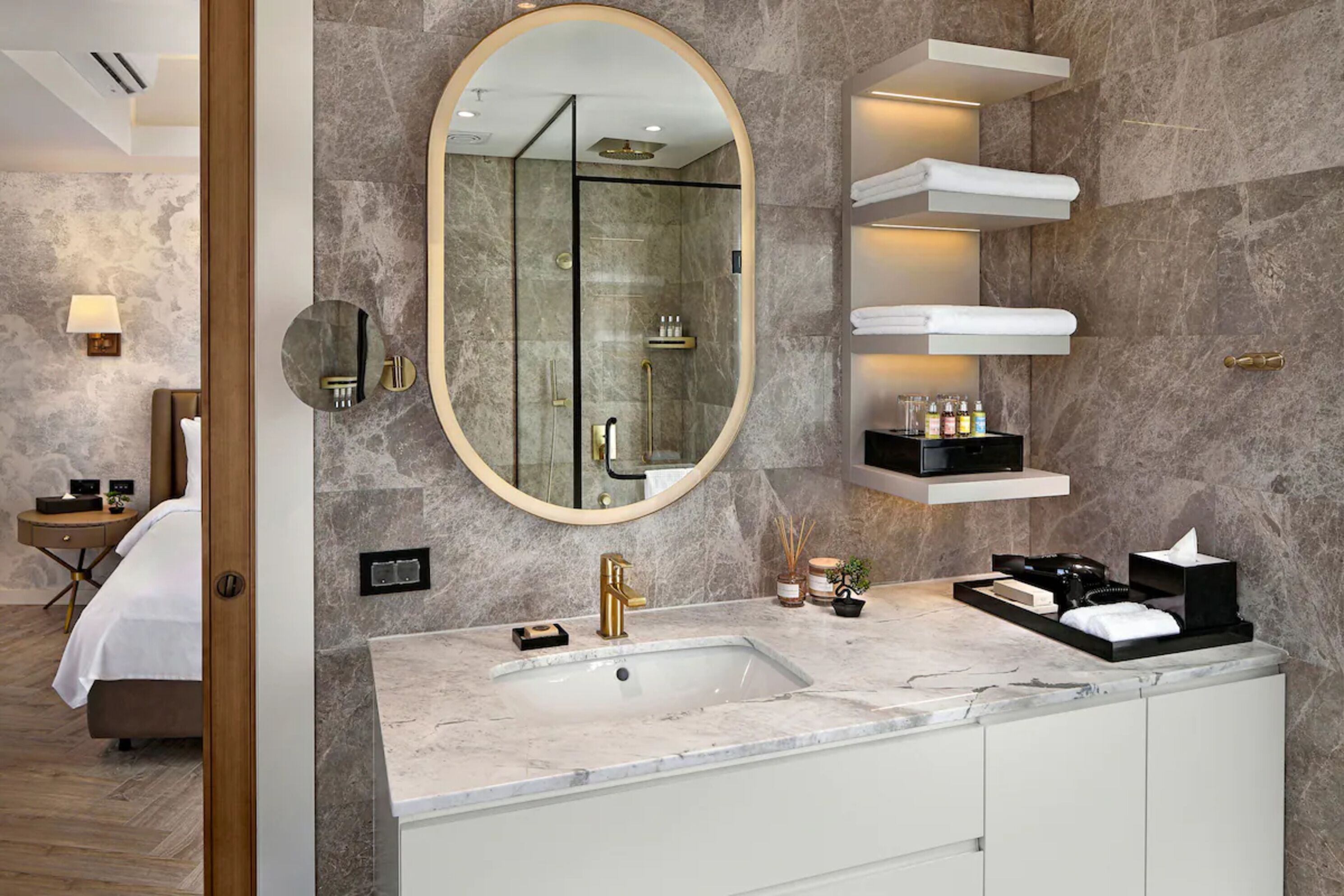 A modern bathroom with a large oval mirror, a gold faucet, and built-in shelves for toiletries.