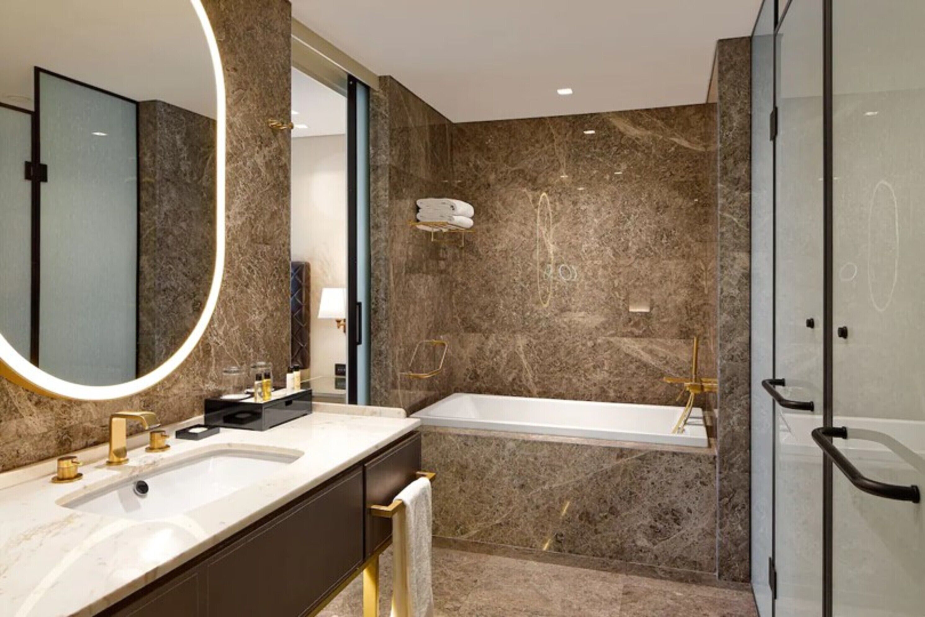 A modern bathroom with a marble wall, a large mirror, a bathtub, and a vanity with a sink.