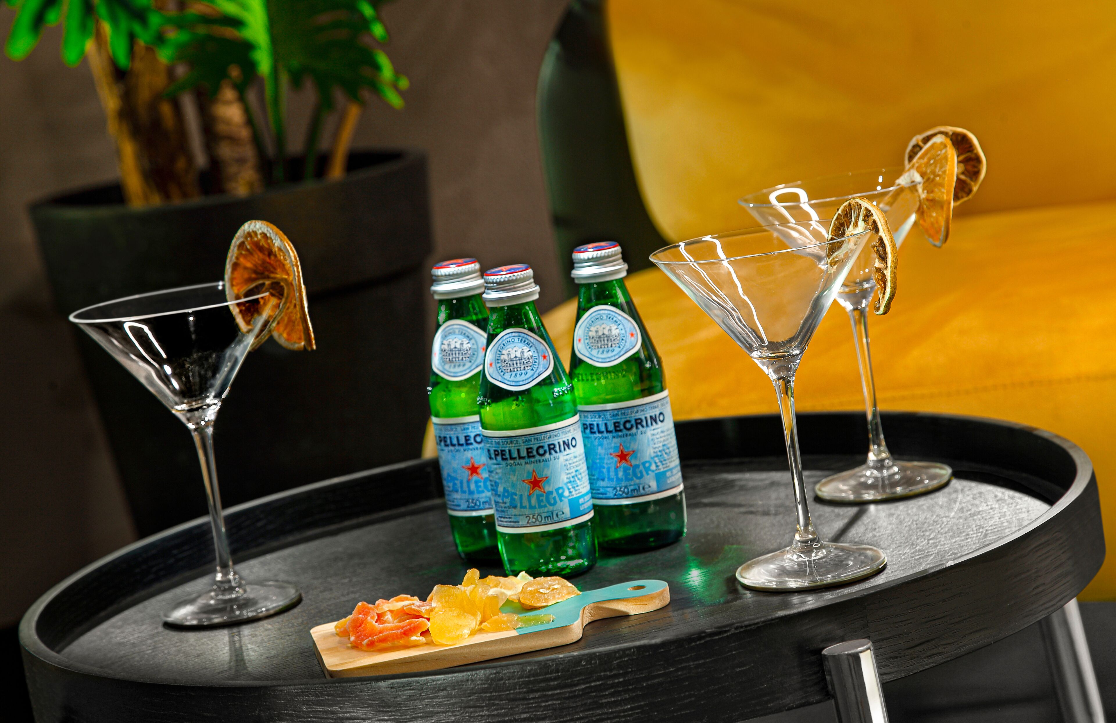 A bar area with three bottles of Pellegrino water, two martini glasses with dried orange slices, and a wooden cutting board with citrus slices.