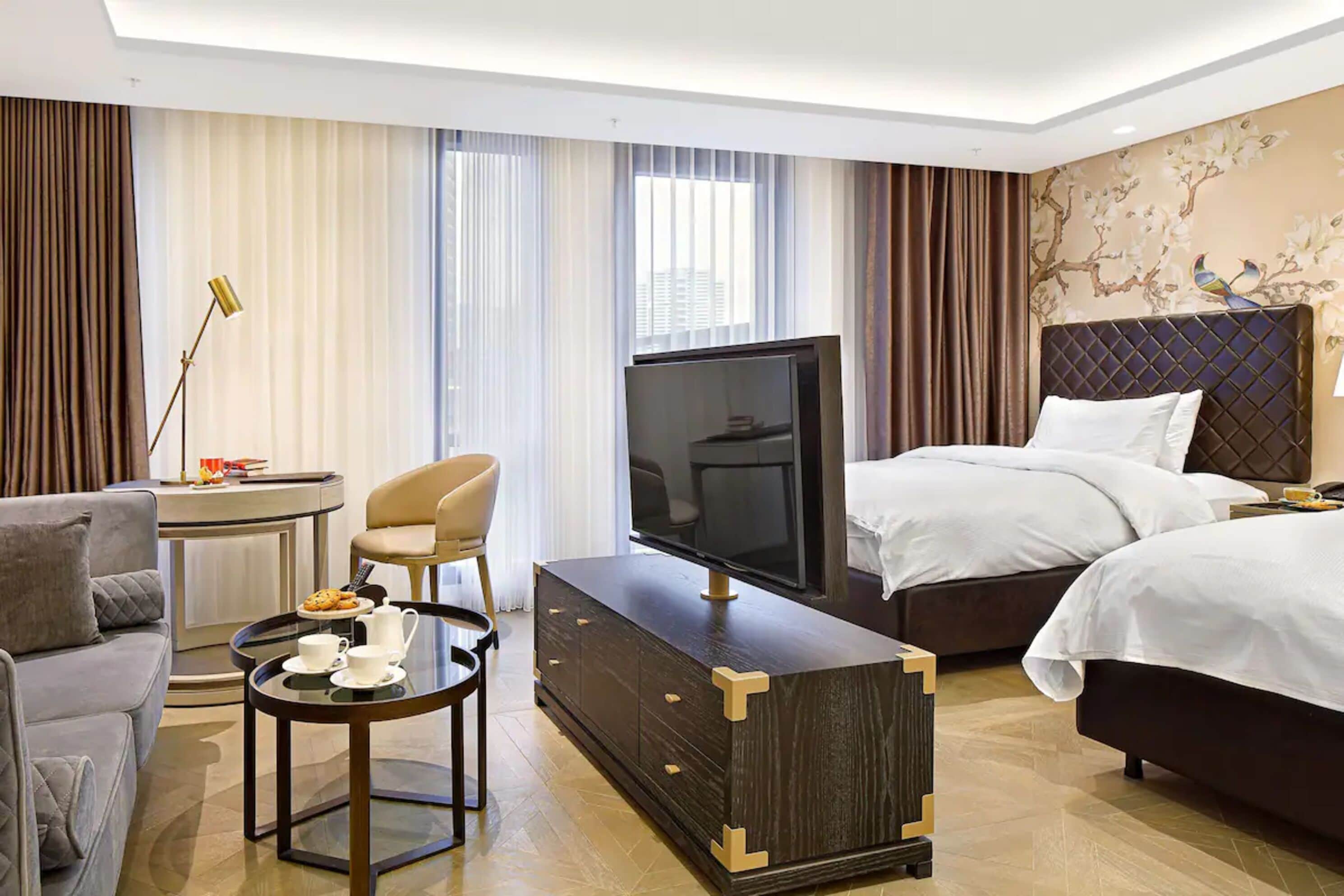 Habitación de hotel con cama, televisión, sofá, sillón, una mesa con una bandeja de comida y un detalle decorativo en la pared.