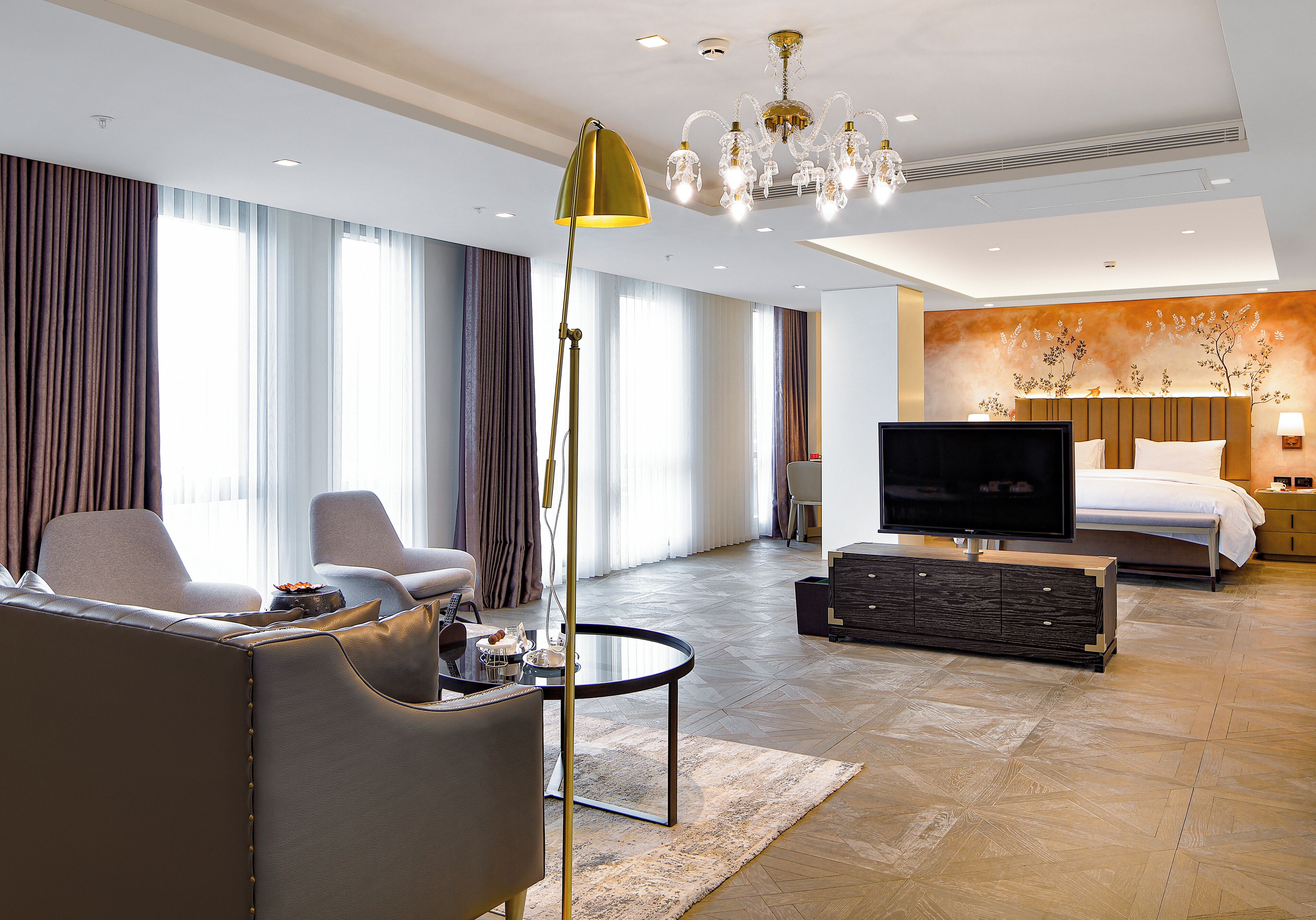 A modern hotel room with a sofa, armchair, TV, bed, and a chandelier.