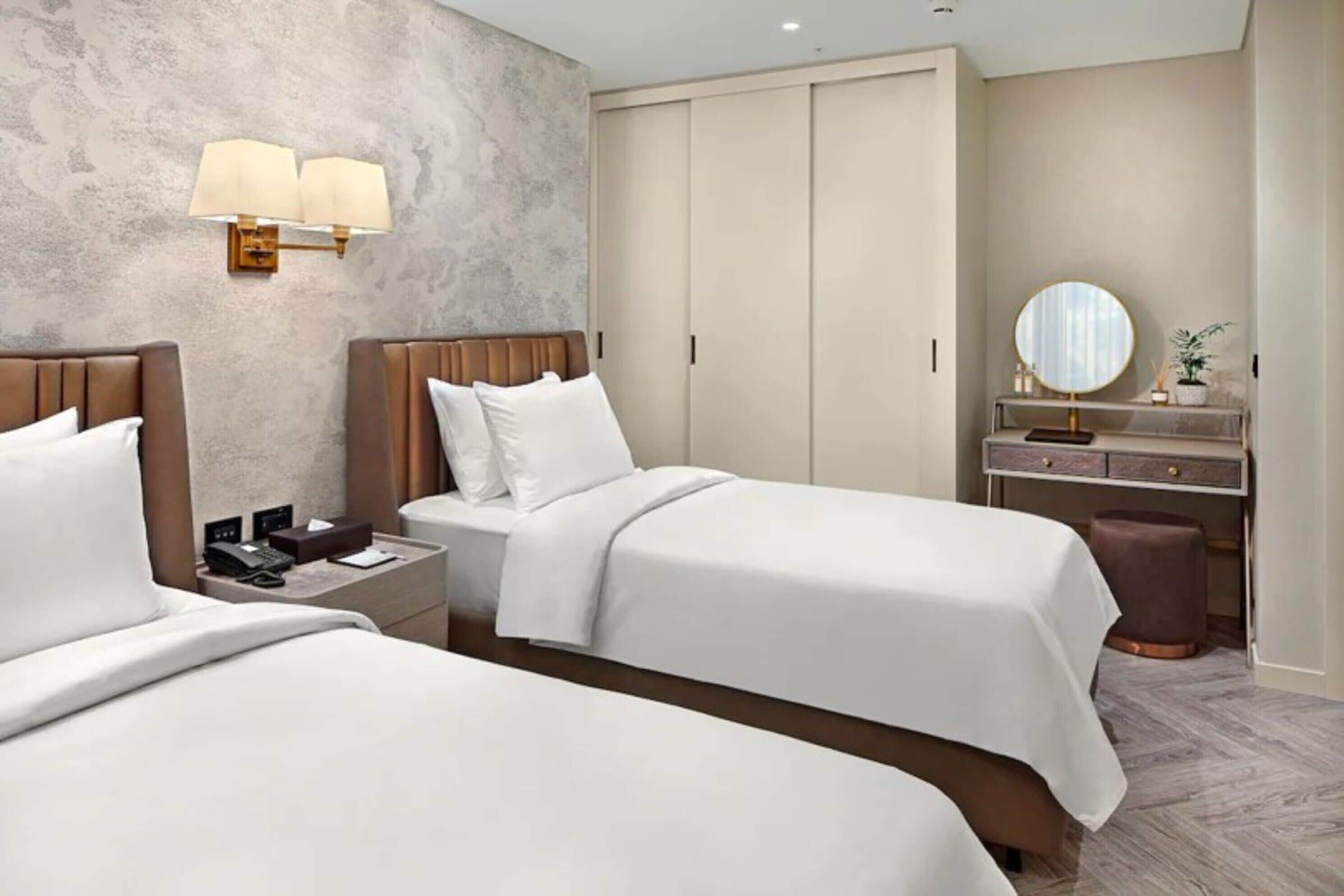Habitación de hotel con dos camas, un armario grande, una tocador con espejo y una pequeña mesa con lámpara.