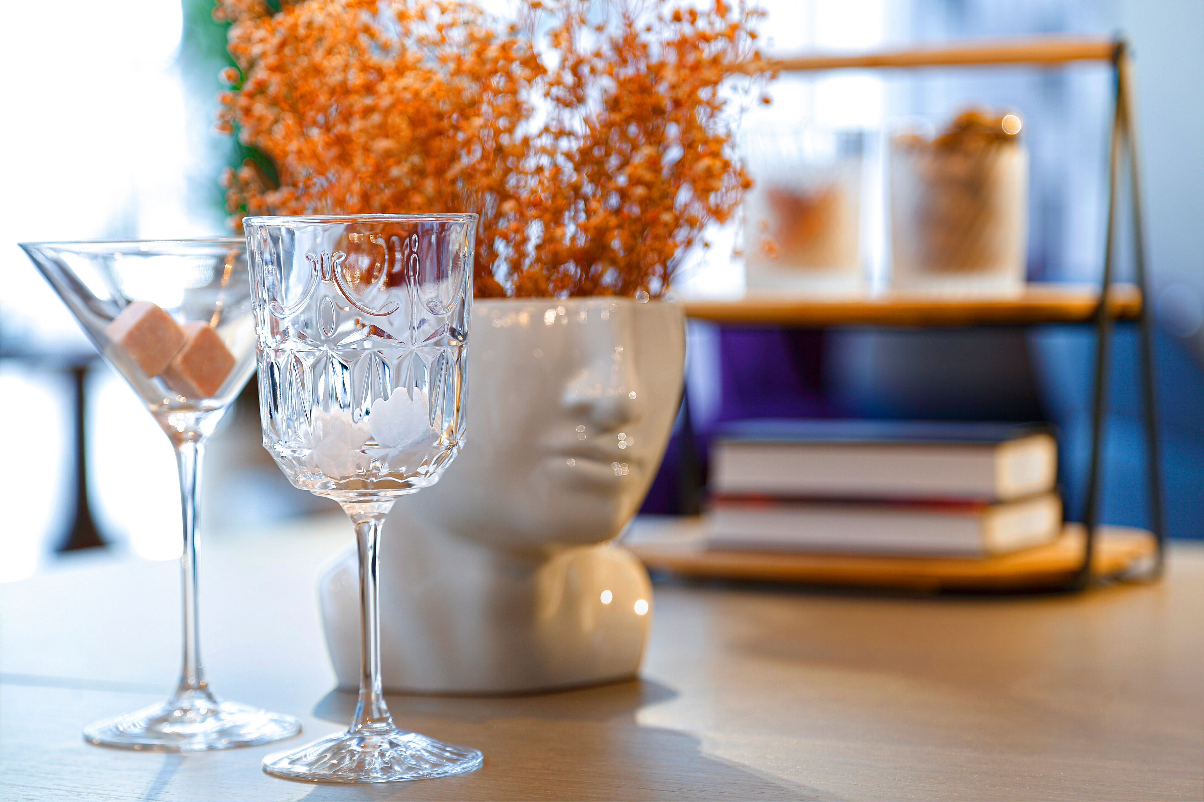 Two clear glasses with ice cubes on a wooden table, a white vase with orange flowers, and a bookshelf in the background.