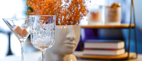 Two clear glasses with ice cubes on a wooden table, a white vase with orange flowers, and a bookshelf in the background.