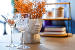 Dois copos de vidro com gelo sobre uma mesa de madeira, um vaso branco com flores laranja e uma estante de livros ao fundo.