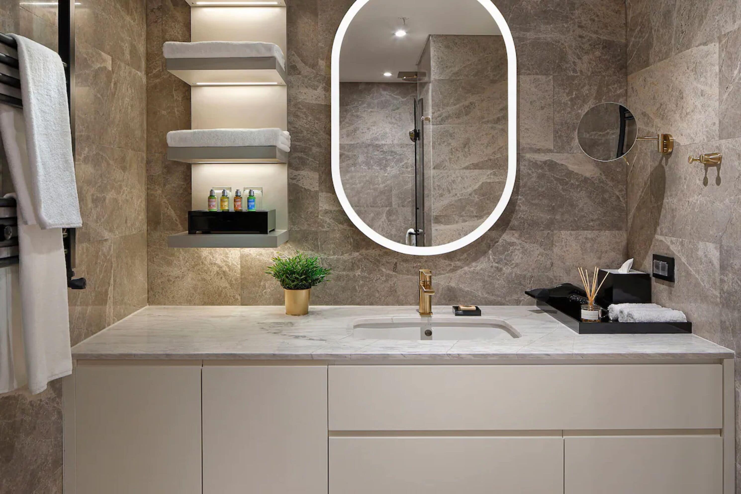 A modern bathroom with a marble countertop, a large oval mirror, a glass-enclosed shower, and a shelf with toiletries.