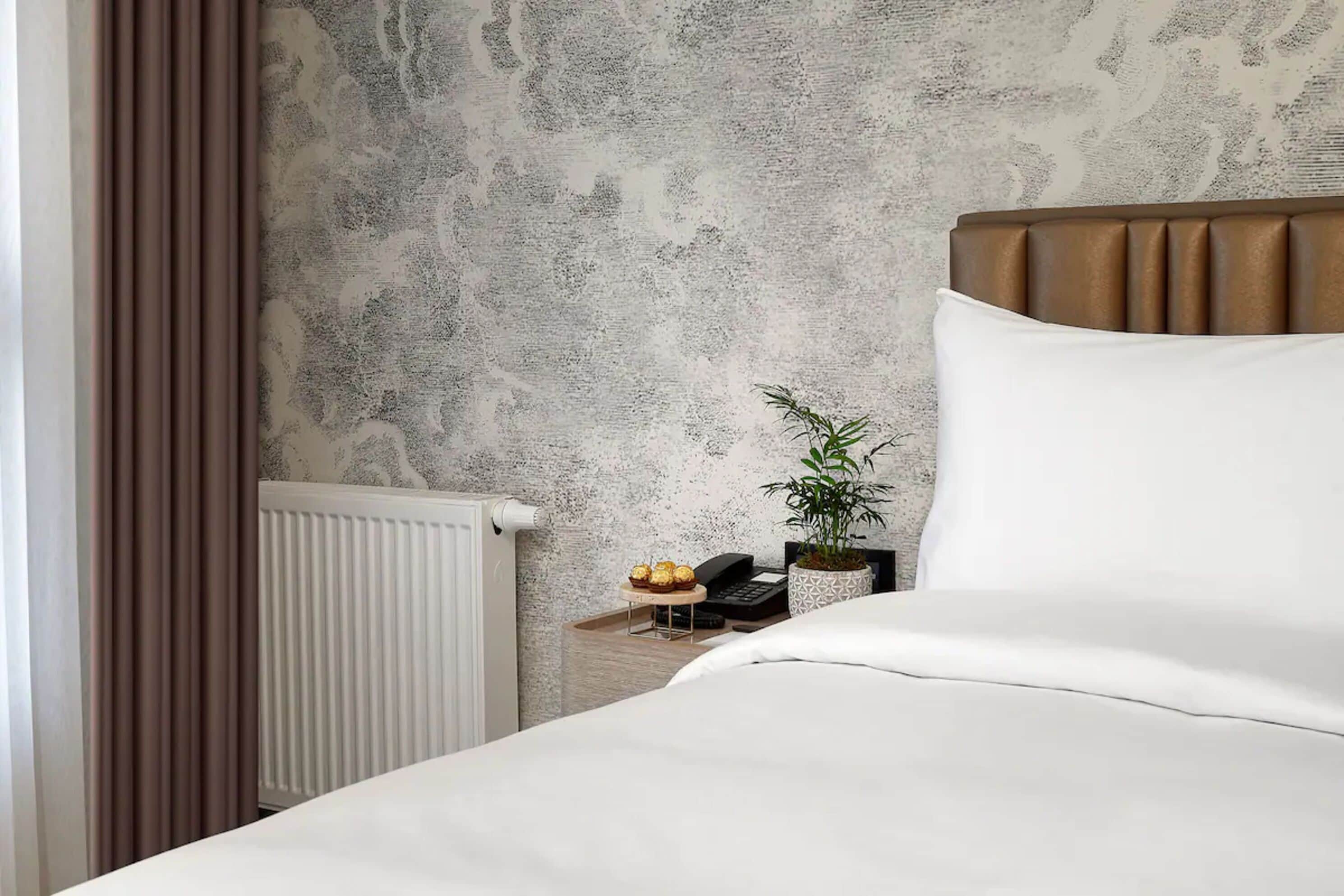 A neatly made bed with white linens, a brown headboard, and a small bedside table with a plant and a phone.