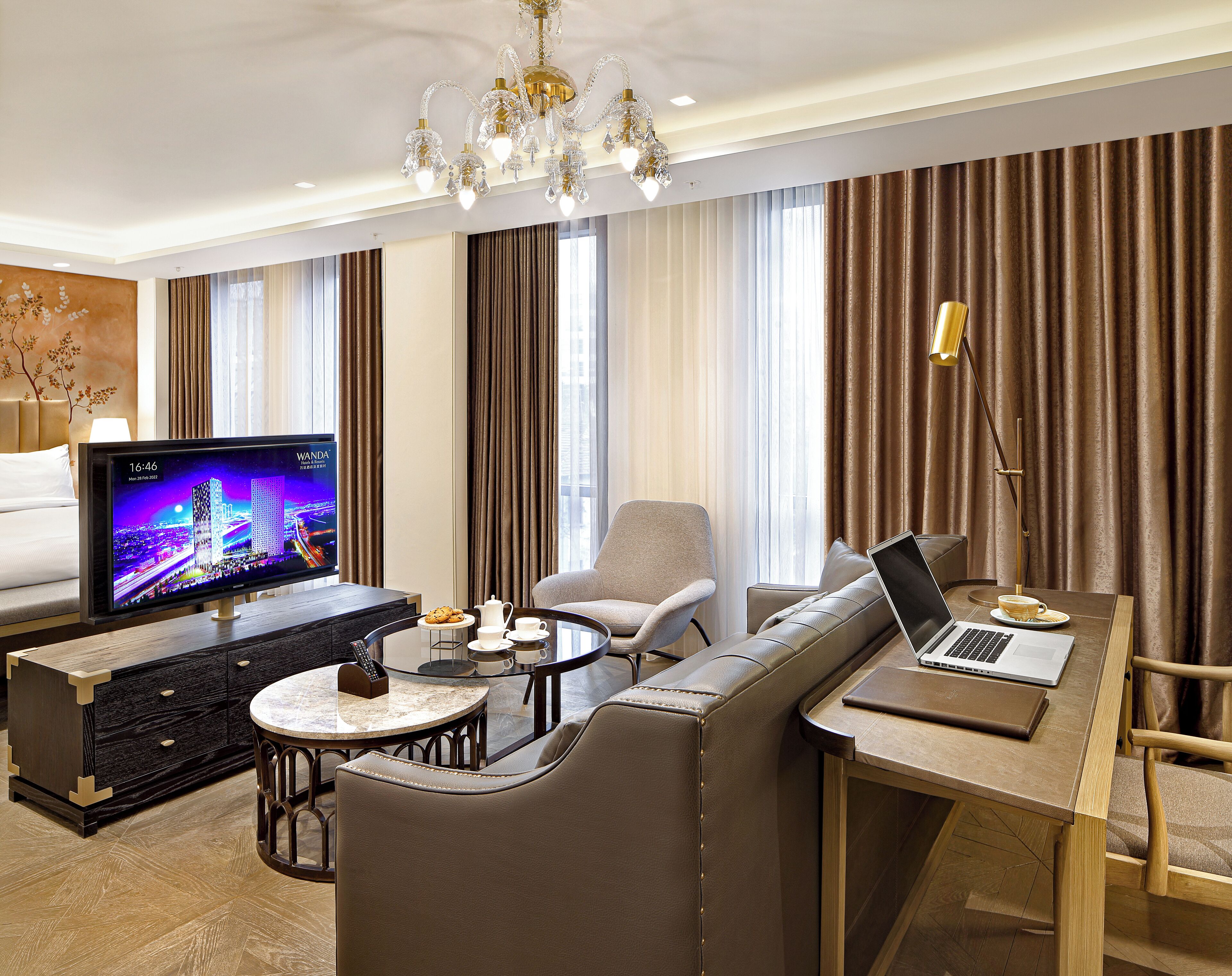 A modern hotel room with a large TV, a sofa, a dining table, and a laptop.