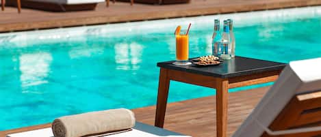 Escena junto a la piscina con sillones de descanso, una mesa con una toalla, un vaso de jugo de naranja y botellas de agua.