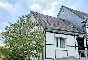 A two-story house with a gabled roof, a visible window, and a tree in front.