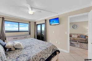 Un dormitorio con cama, ventilador de techo, televisión, reloj y vista al océano.