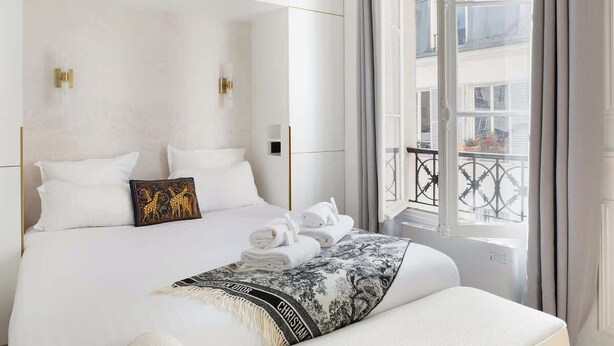 A hotel room with a large bed, white bedding, a decorative throw, and a view of the city through a balcony. - A Dazzling 2-BR apartment in Le Marais (Paris)