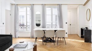 A modern living room with a dining area, large windows with curtains, a black sofa, a white table, and a black fireplace. - A Dazzling 2-BR apartment in Le Marais (Paris)