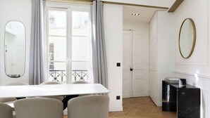 A modern room with a white dining table, four chairs, a black marble side table, and a large window with curtains. - A Dazzling 2-BR apartment in Le Marais (Paris)