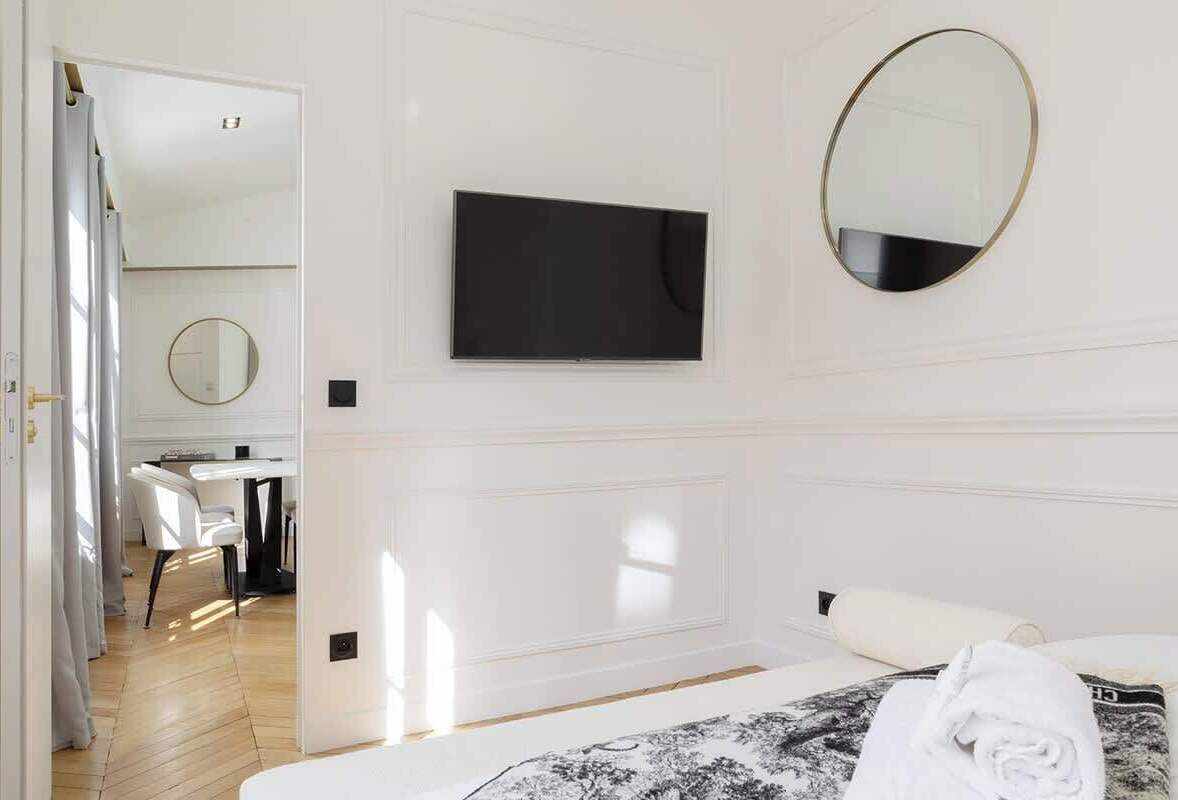 A modern hotel room with a bed, a flat-screen TV, a round mirror, and a dining area visible through an open door.
