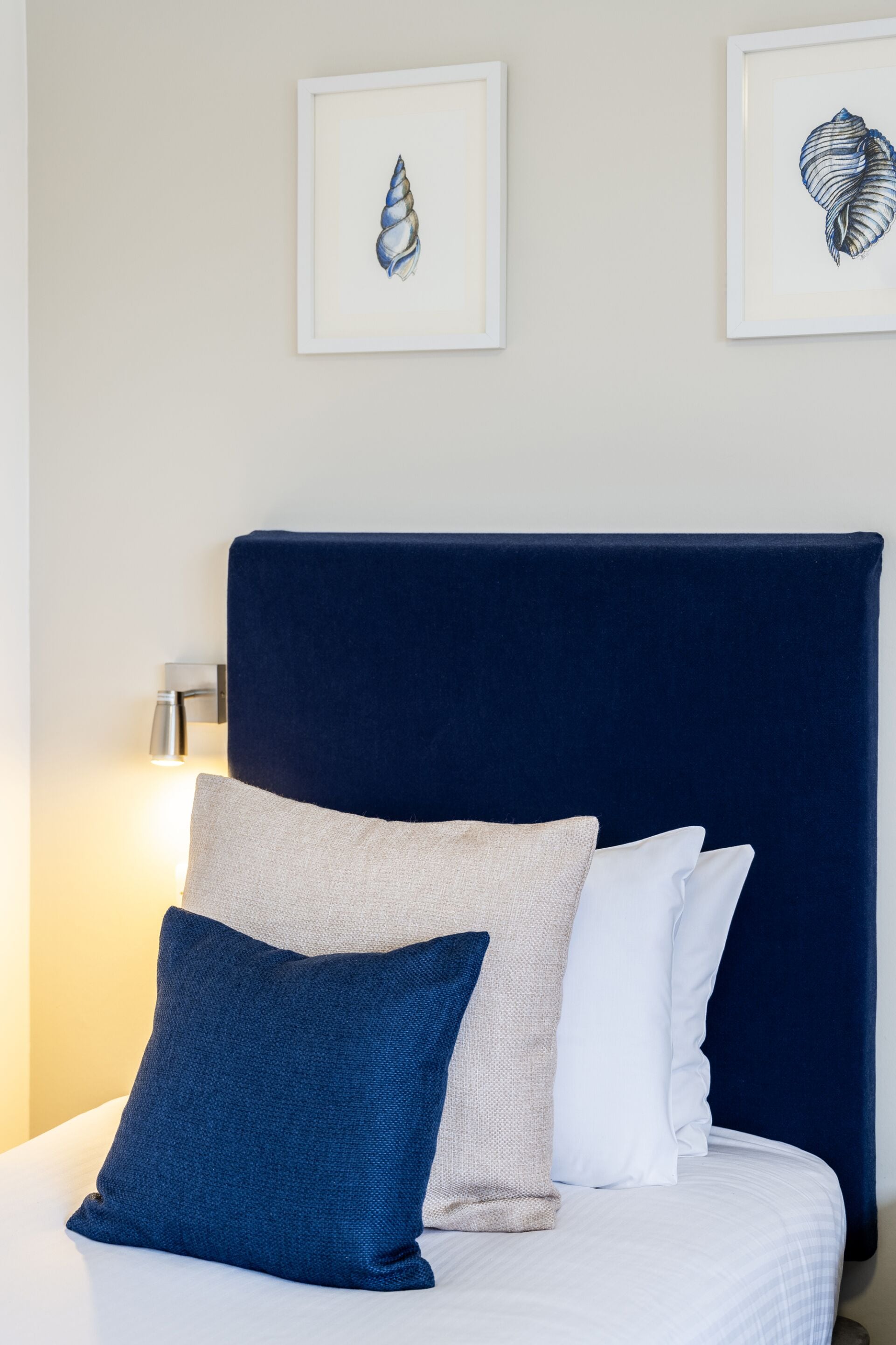 Ein ordentlich bezogenes Bett mit dunklem Kopfteil, einer Nachttischlampe und zwei gerahmten Kunstwerken an der Wand.