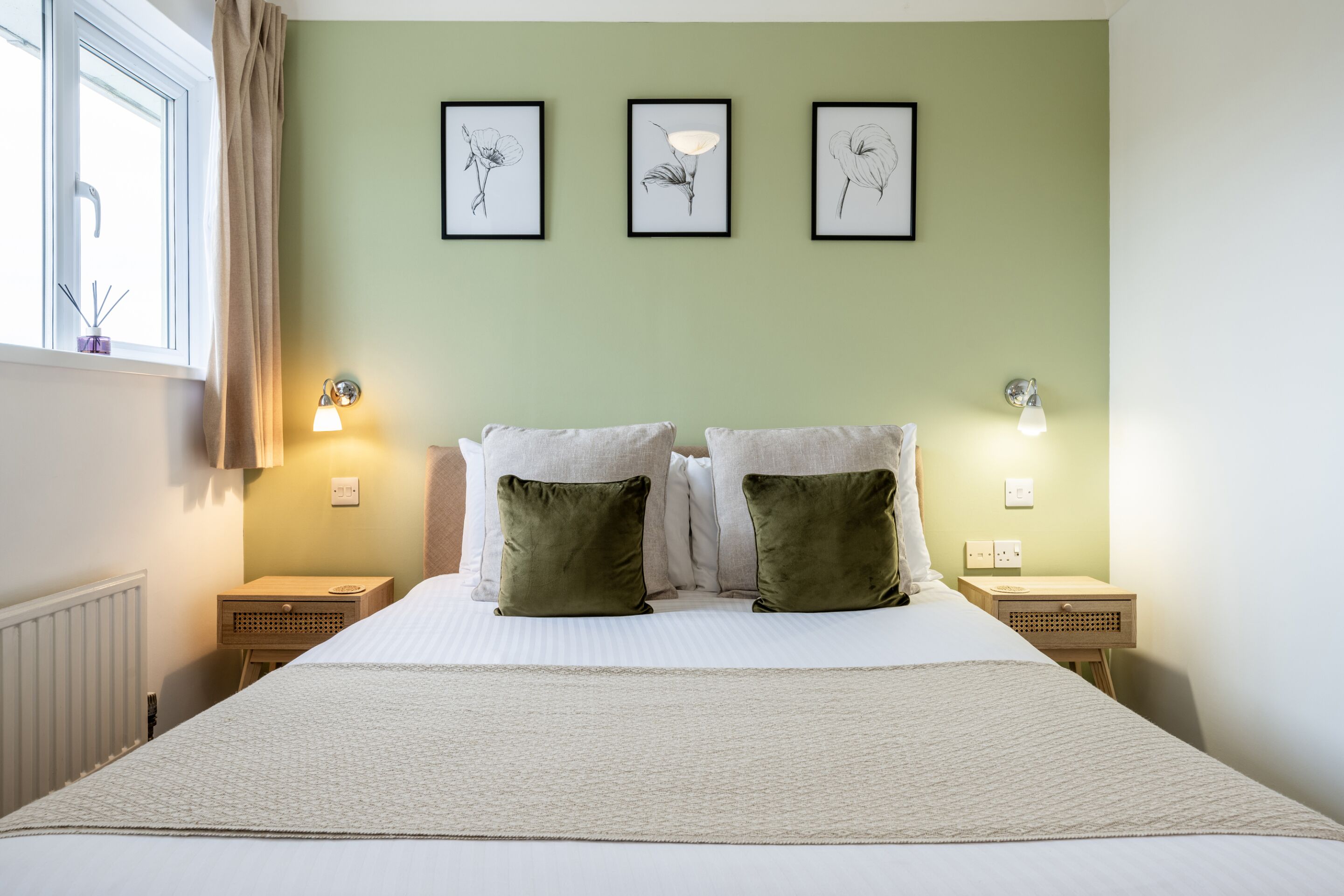 A neatly made bed with two pillows, a bedside table with a lamp, and three framed botanical prints on the wall.