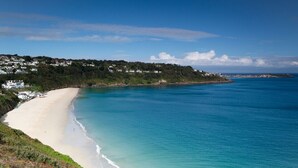 A coastal landscape with a sandy beach, clear blue waters, and a hillside with buildings.