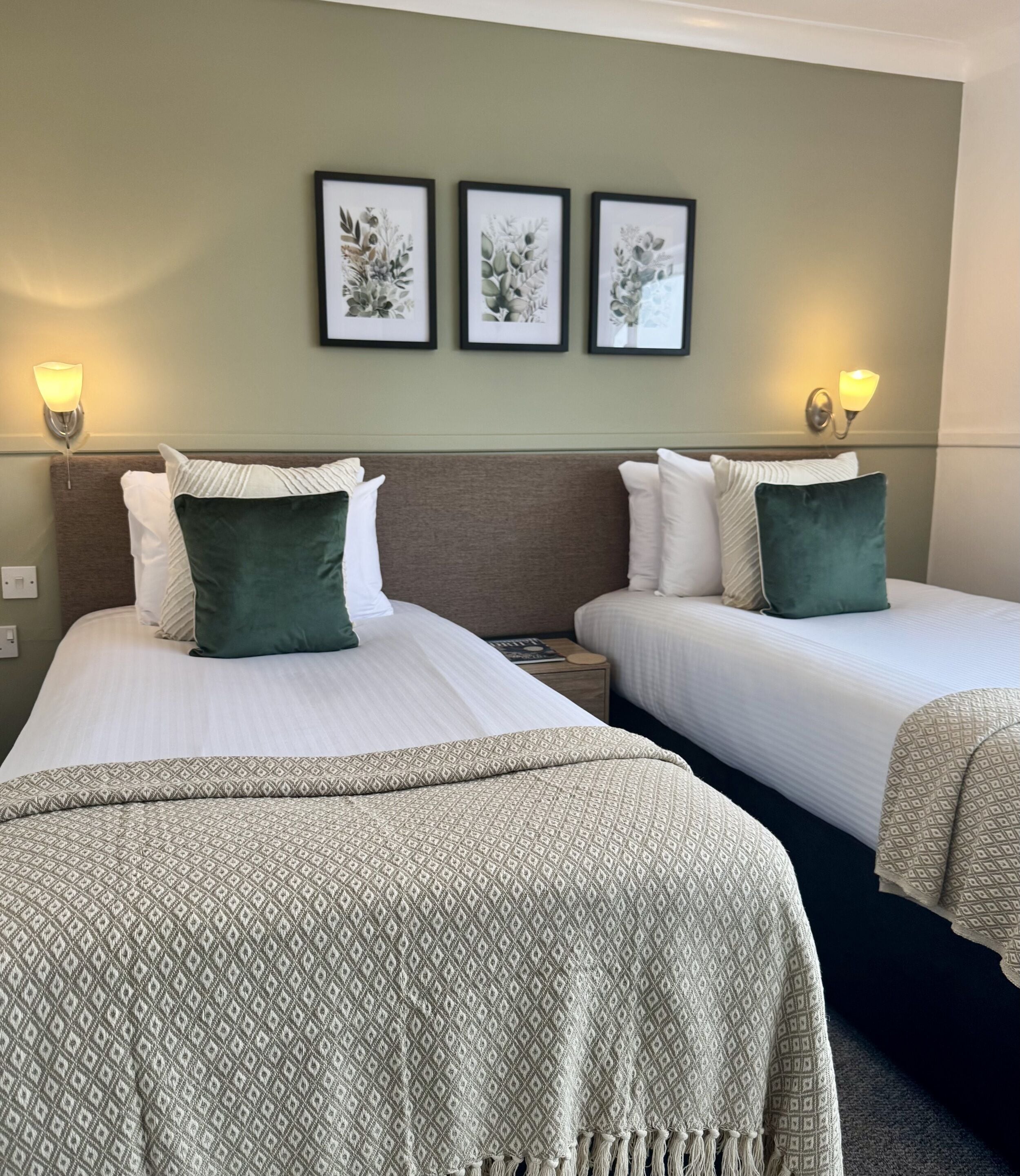 Ett hotellrum med två sängar, var och en med ett mönstrat sängöverkast, en sänggavel och ett nattduksbord. På väggen ovanför sängarna finns väggmonterade lampor och inramade botaniska illustrationer.
