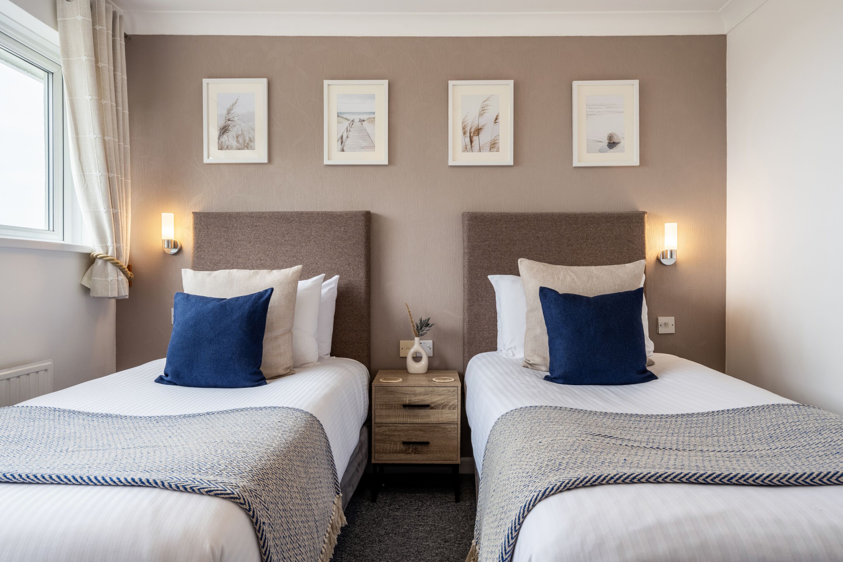 A double room with two beds, each with a headboard, bedside tables, and wall-mounted lamps. There are four framed pictures on the wall above the beds.
