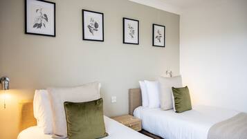 A hotel room with two beds, a nightstand, a wall-mounted lamp, and framed botanical prints on the wall.