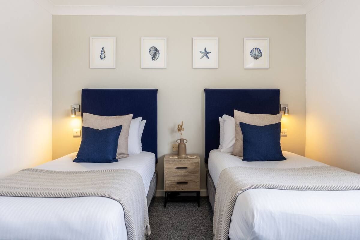 Ein Hotelzimmer mit zwei Betten, jeweils mit Kopfteil, Nachttischen mit Lampen und Wandbildern.