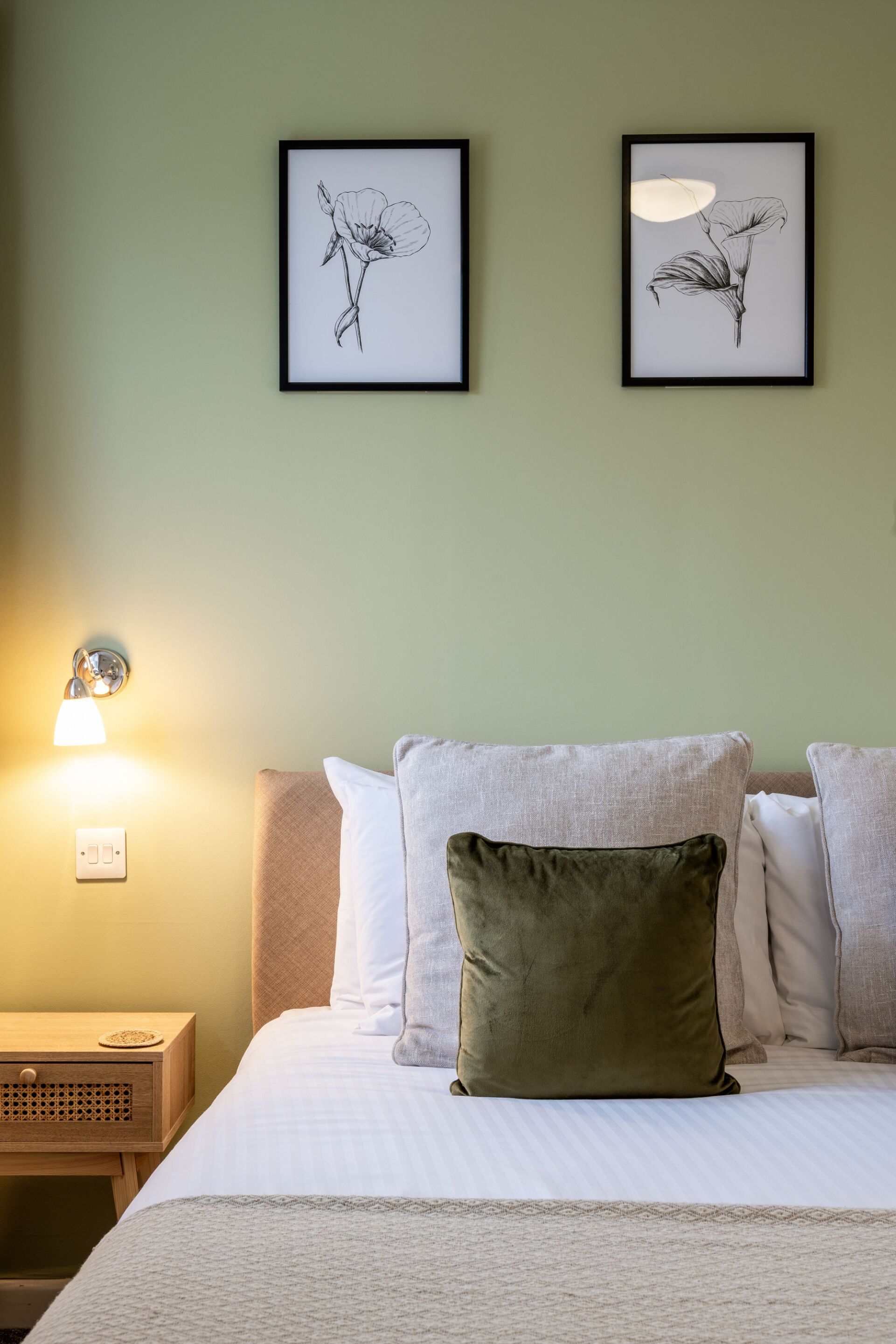 Een netjes opgemaakt bed met een witte dekbedovertrek en een groen kussen, een nachtkastje met een lamp, en twee ingelijste botanische prints aan de muur.
