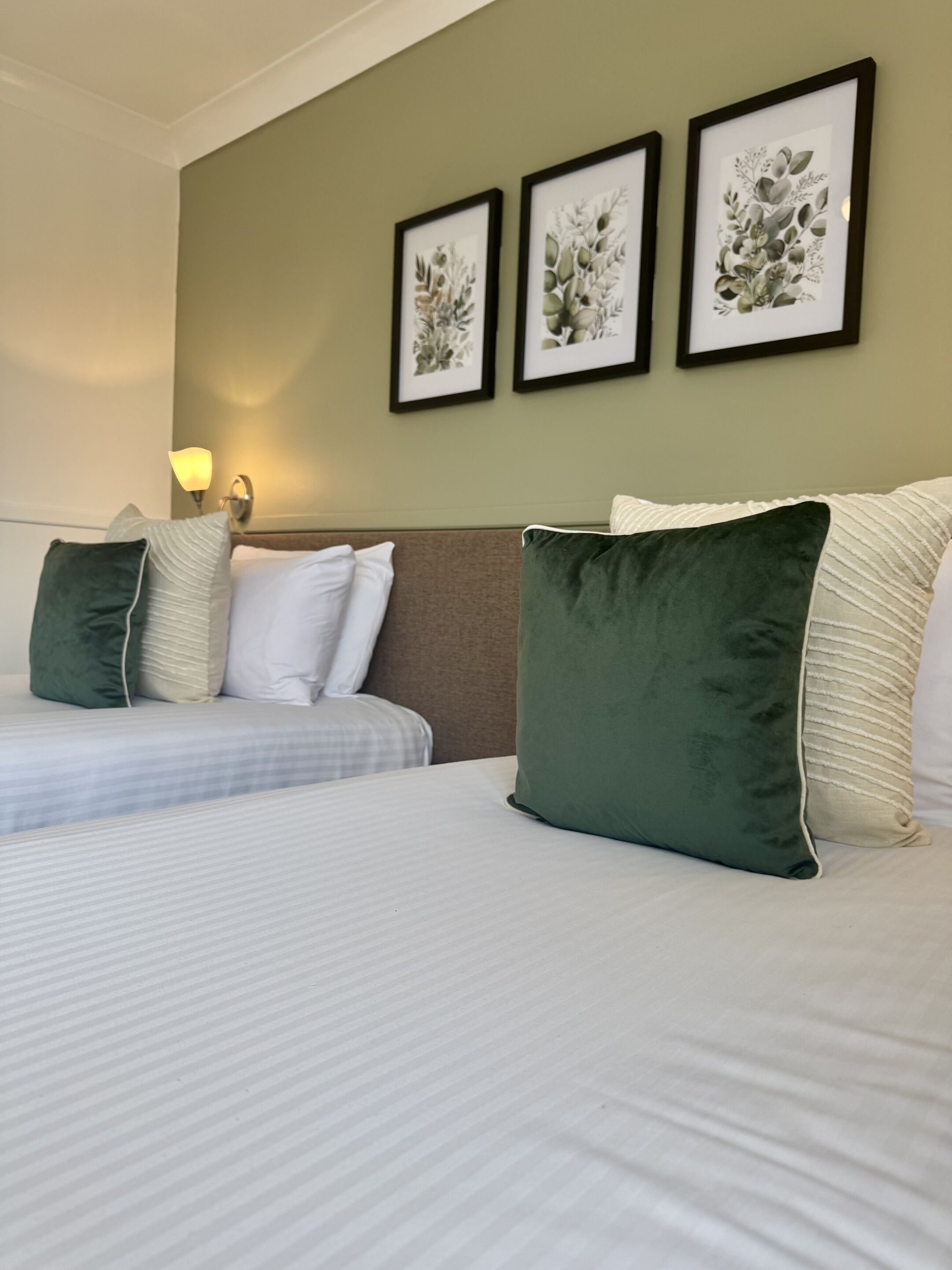 Habitación de hotel con dos camas, almohadas verdes y blancas, y una obra de arte botánica enmarcada en la pared.