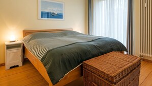 Un lit bien fait, recouvert d’une couvre-lit à rayures, une table de chevet avec une lampe, un banc en osier et une fenêtre avec des rideaux.