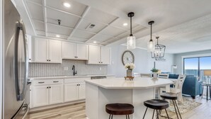 A modern kitchen with white cabinetry, a central island with stools, pendant lighting, and a view to the living area.