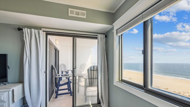 A room with a large window, a sliding glass door leading to a balcony with chairs, a bed, and a TV.