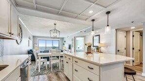 A modern kitchen with white cabinetry, a central island, pendant lighting, and a view to the living area.