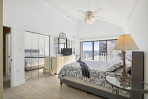A bedroom with a large bed, a television, a ceiling fan, and a balcony with a view of the sea.