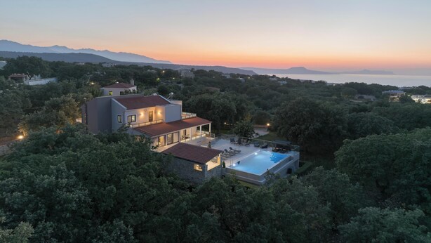 Une maison moderne avec une piscine, entourée d’une végétation luxuriante, et dont le décor est constitué de montagnes et d’un coucher de soleil.