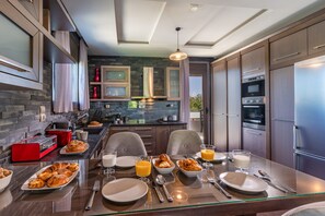 A modern kitchen with a dining area, featuring a stone backsplash, stainless steel appliances, and a glass dining table set for breakfast.