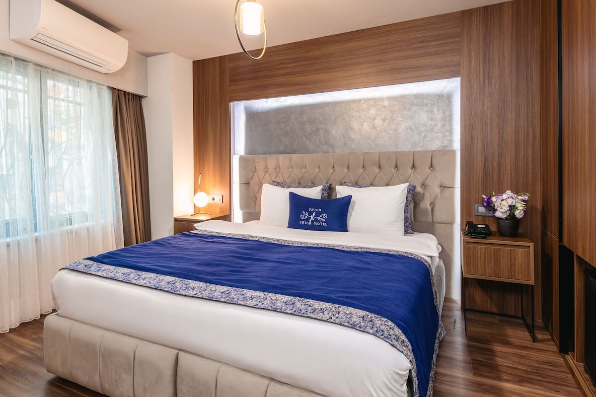 Ein Hotelzimmer mit einem großen Bett, Holzvertäfelung, einem Nachttisch mit Lampe und Blick ins Freie durch ein Fenster.