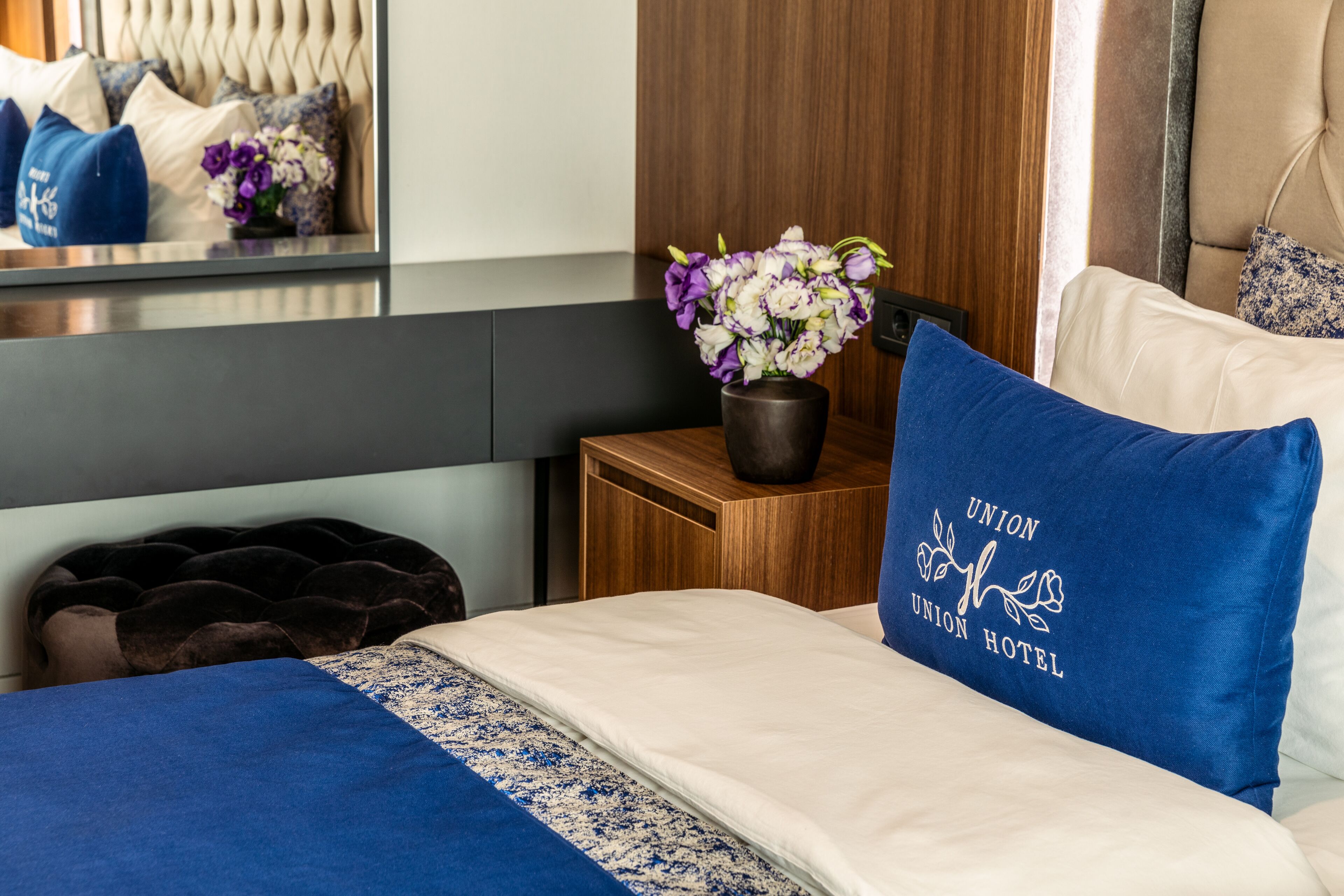Habitación de hotel con una cama, una mesita de noche con un florero y un almohadón azul con el nombre del hotel.
