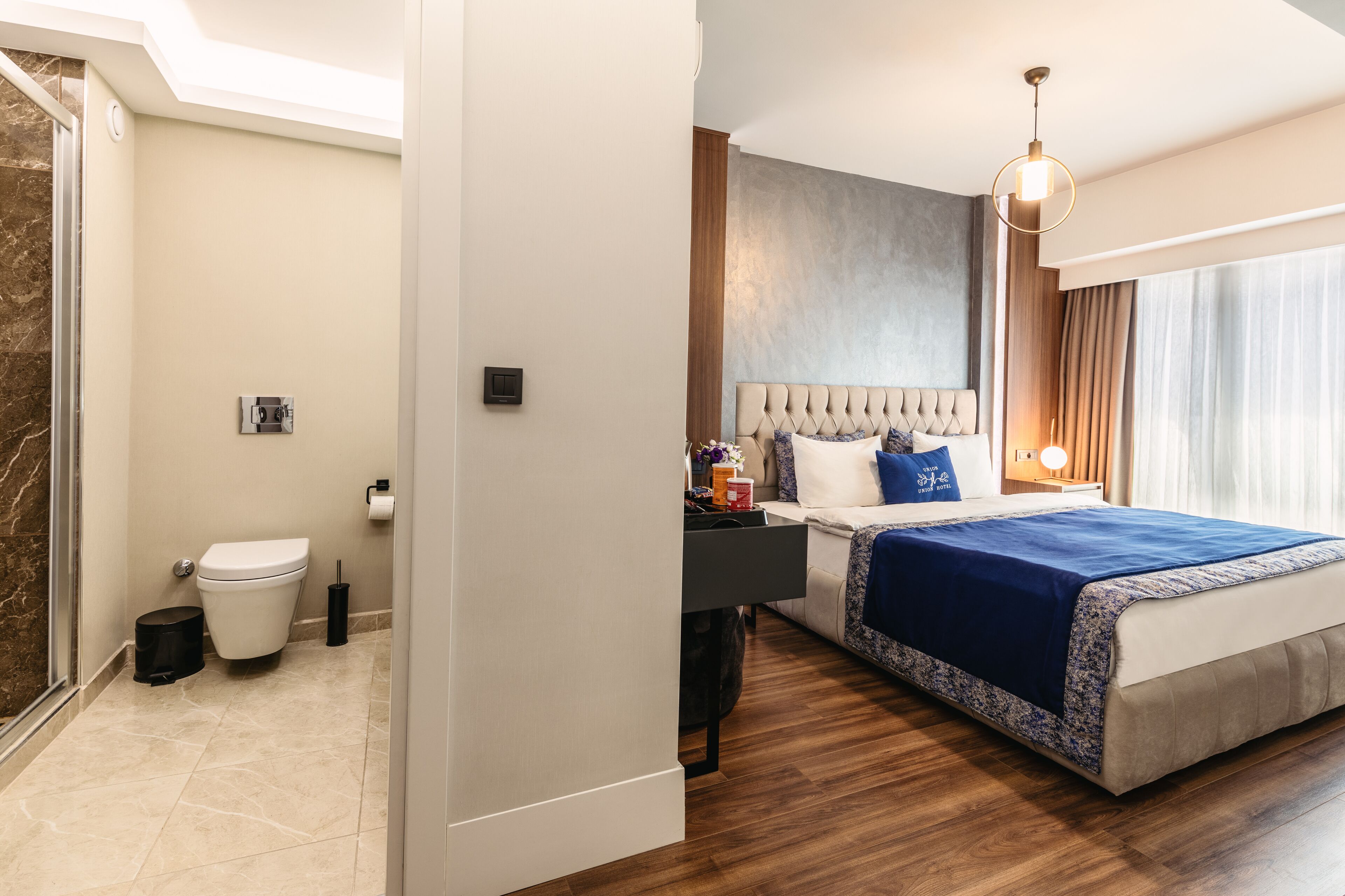 A modern hotel room with a large bed, a bedside table, a nightstand, a bathroom with a toilet, and a shower area.