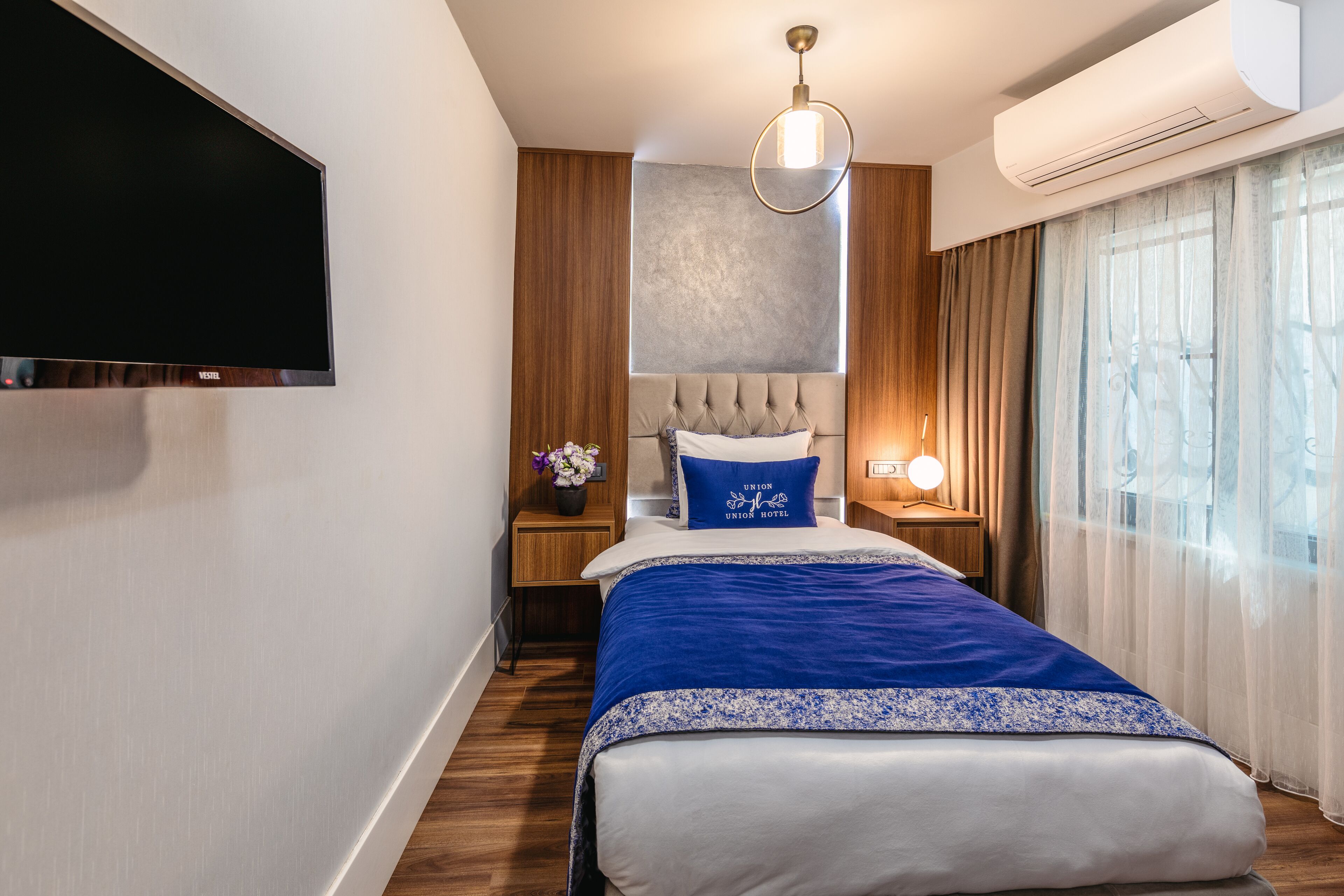 A modern hotel room with a large bed, a flat-screen TV, a wall-mounted air conditioner, and a window with sheer curtains.