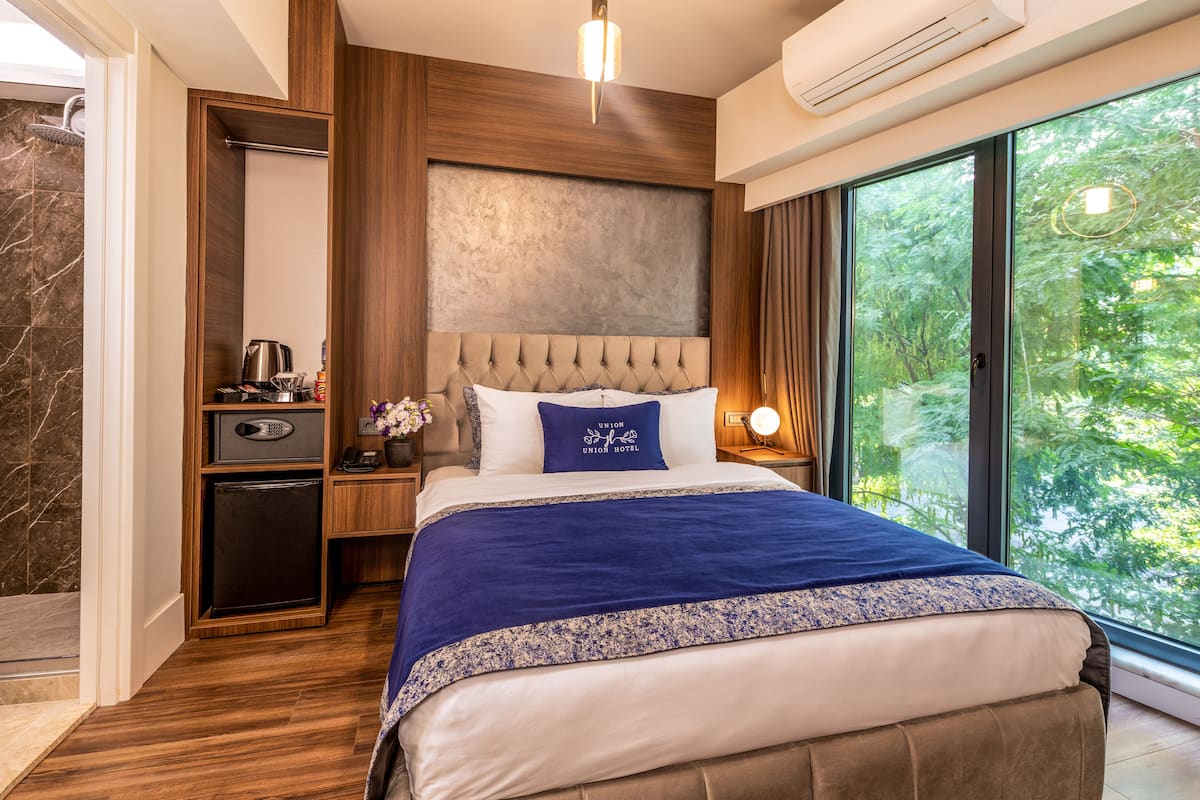 Ein modernes Hotelzimmer mit einem großen Bett, einem eingebauten Kleiderschrank, einer kleinen Küchenzeile und einem großen Fenster mit Blick auf Bäume.