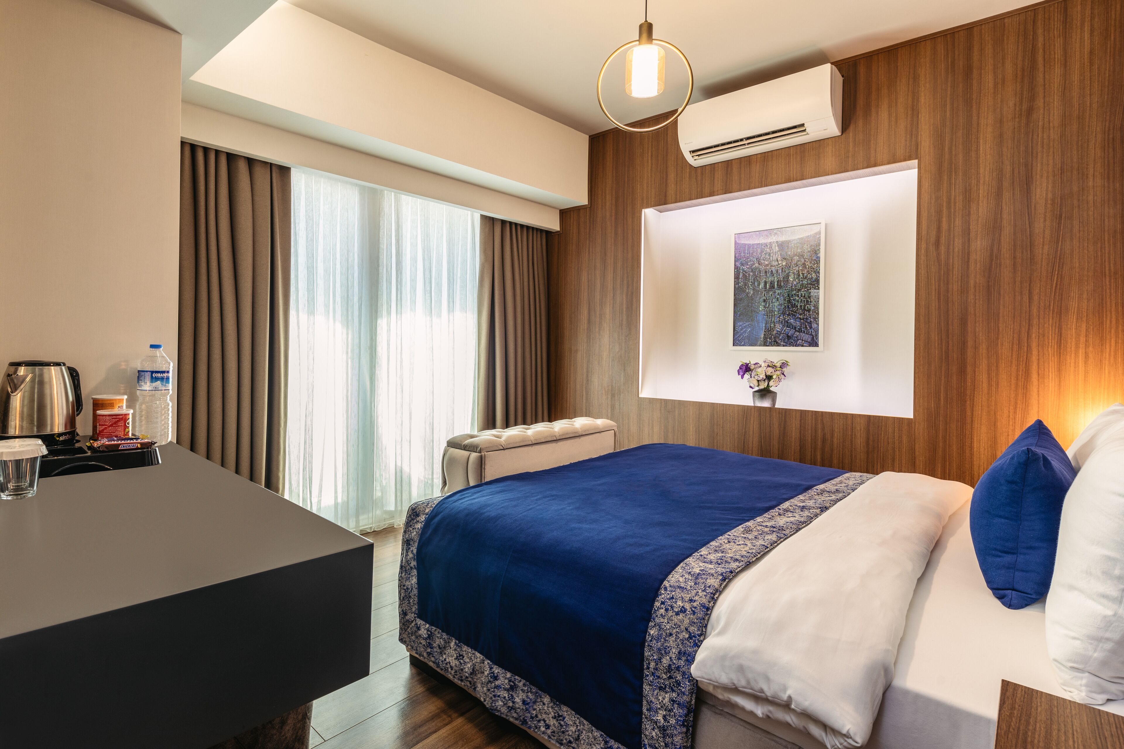 A hotel room with a bed, a desk, a chair, a window with curtains, an air conditioning unit, and a wall-mounted picture.