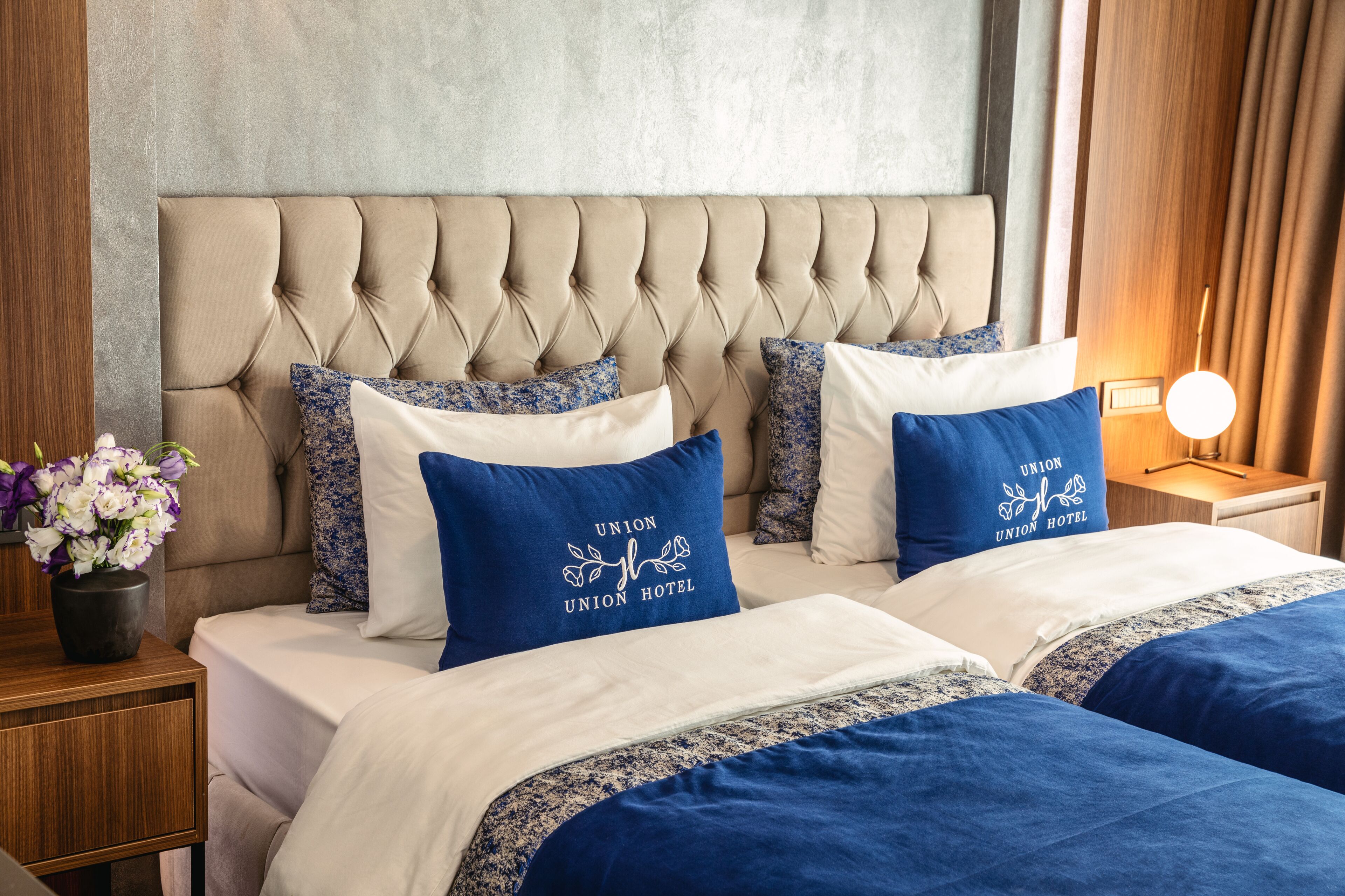 A hotel room with a tufted headboard, two beds with blue and white bedding, and a nightstand with a flower vase.