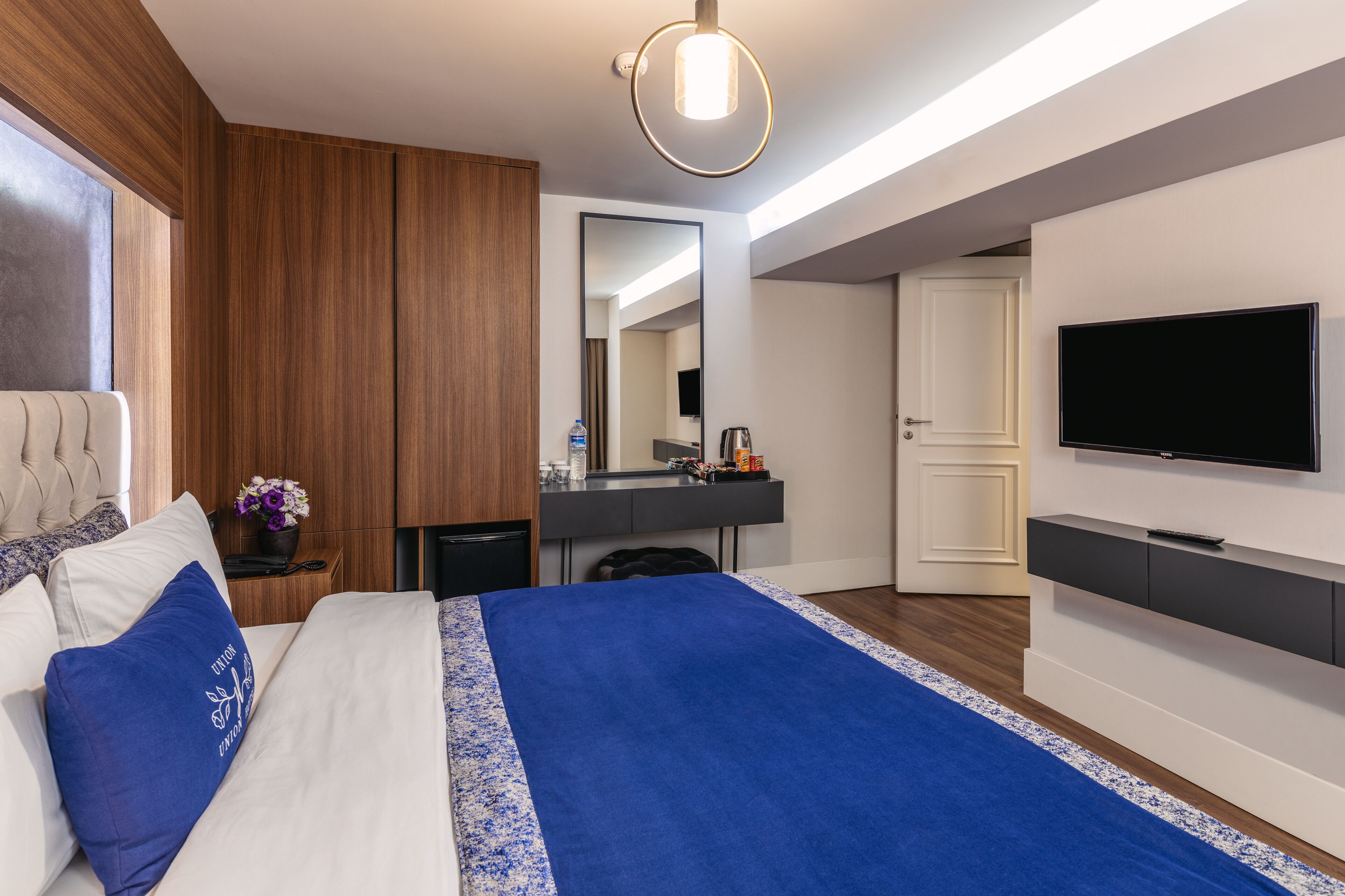 A modern hotel room with a large bed, a flat-screen TV, a wooden panel wall, a small desk with a mirror, and a bathroom door.
