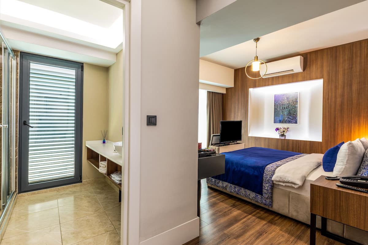 Ein modernes Hotelzimmer mit Bett, Nachttisch, Fernseher und einem großen Fenster mit Jalousien.