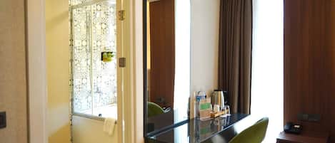 A hotel room with a glass shower enclosure, a black desk with a chair, a mirror, a wooden cabinet, and a window with curtains.