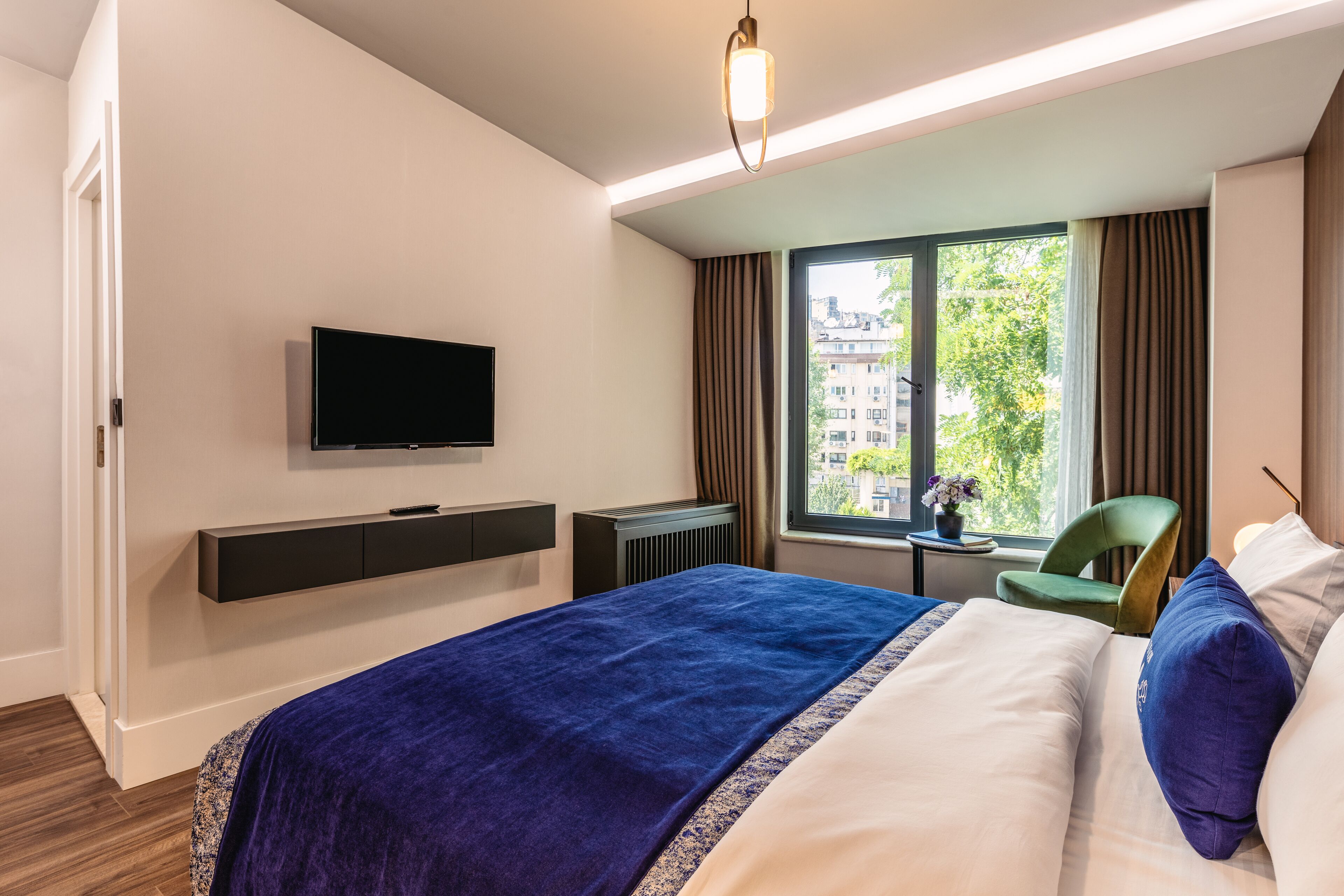 A hotel room with a large bed, a flat-screen TV mounted on the wall, a window with curtains, a green chair, and a small table with a vase.
